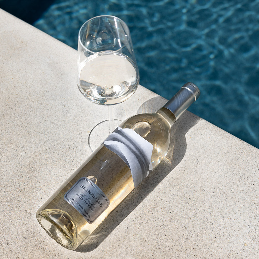 A bottle of Blindfold Blanc de Noir White Blend, placed on a light stone surface beside a clear wine glass, with blue pool water in the background.