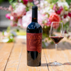 A bottle of Domain Curry Napa Valley Cabernet Sauvignon stands on a wooden table beside a filled wine glass, with a blurred floral background.