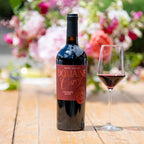 A bottle of Domaine Curry Napa Valley red wine stands on a wooden table beside a filled wine glass, with a blurred floral background.