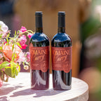 Two bottles of Domaine Curry Napa Valley red wine standing on a round wooden table, with pink and peach flowers blurred in the foreground and a warm outdoor background.