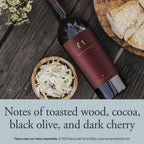 A bottle of Harvey & Harriet red wine lies on a dark wooden table beside a bowl of coleslaw, a plate of bread, and small white flowers. A text banner at the bottom reads: 'Notes of toasted wood, cocoa, black olive, and dark cherry.'