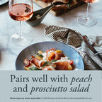 A plated peach and prosciutto salad on a blue plate, styled on a marble table with wine glasses, a striped napkin, and cutlery. Text overlay reads 'Pairs well with peach and prosciutto salad' with small wine branding at the bottom.