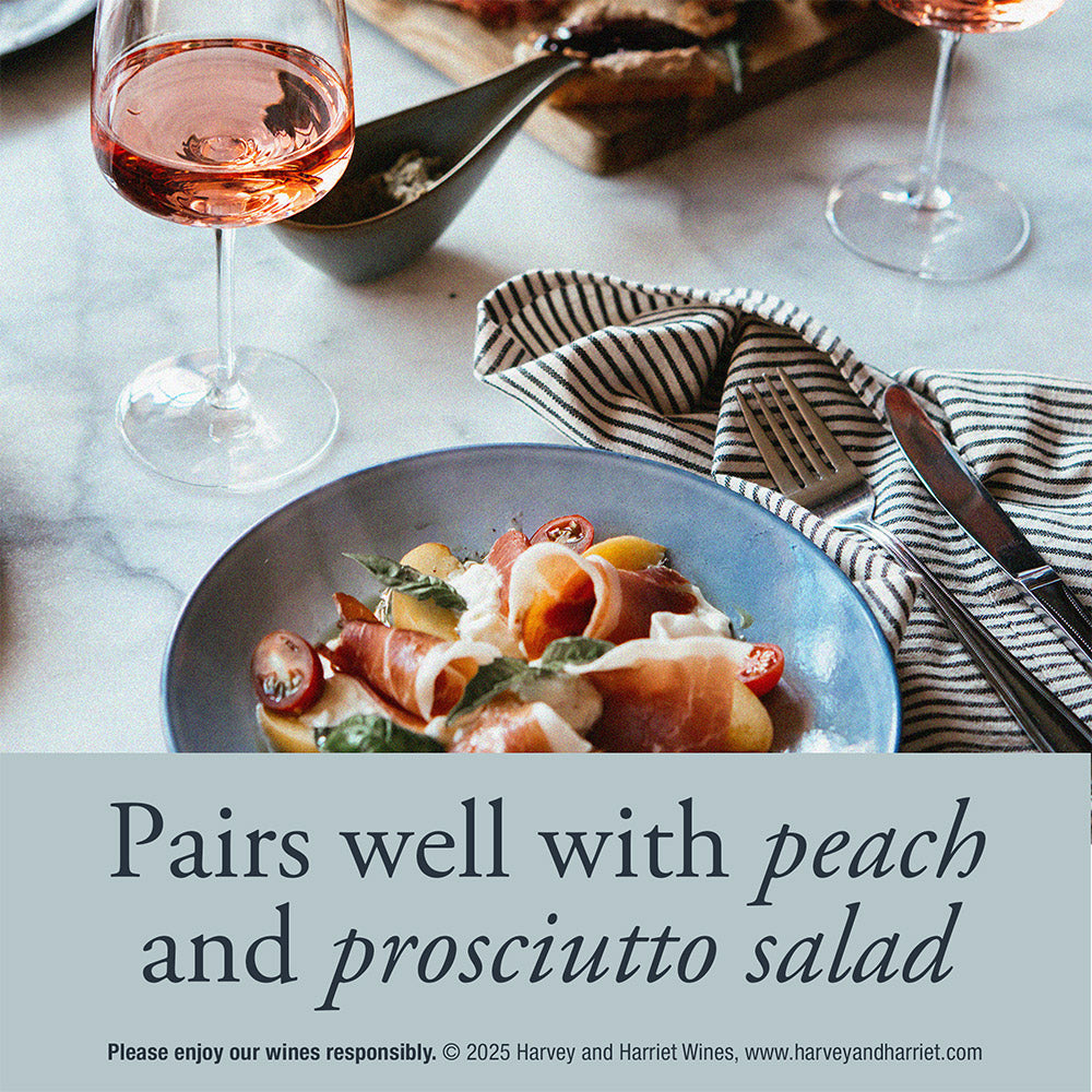 A plated peach and prosciutto salad on a blue plate, styled on a marble table with wine glasses, a striped napkin, and cutlery. Text overlay reads 'Pairs well with peach and prosciutto salad' with small wine branding at the bottom.