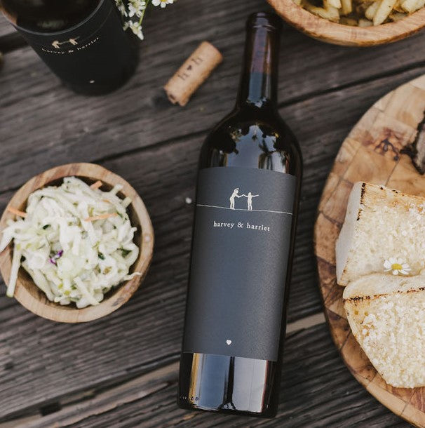 A bottle of 2021 Harvey & Harriet Red Blend laying label side up on a rustic table with plates of food surrounding it.