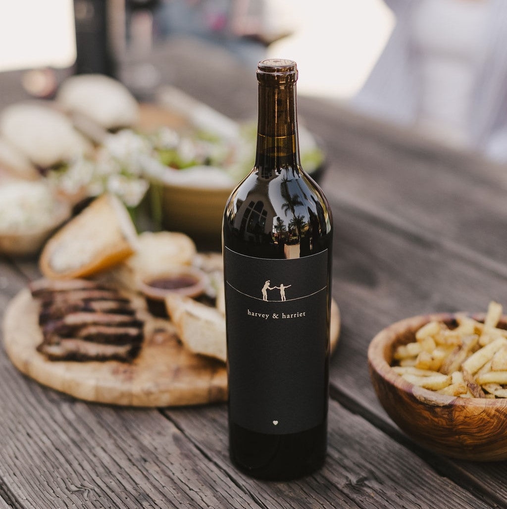 A bottle of 2021 Harvey & Harriet Red Blend on a rustic table with plates of food surrounding it.