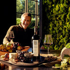 A man at a table with cheese, fruit, a bottle of Mount Veeder Winery Estate Merlot, and a moss wall.