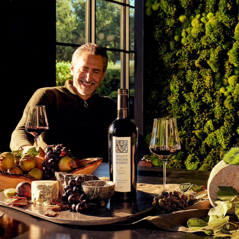 A man at a table with cheese, fruit, a bottle of Mount Veeder Winery Estate Merlot, and a moss wall.
