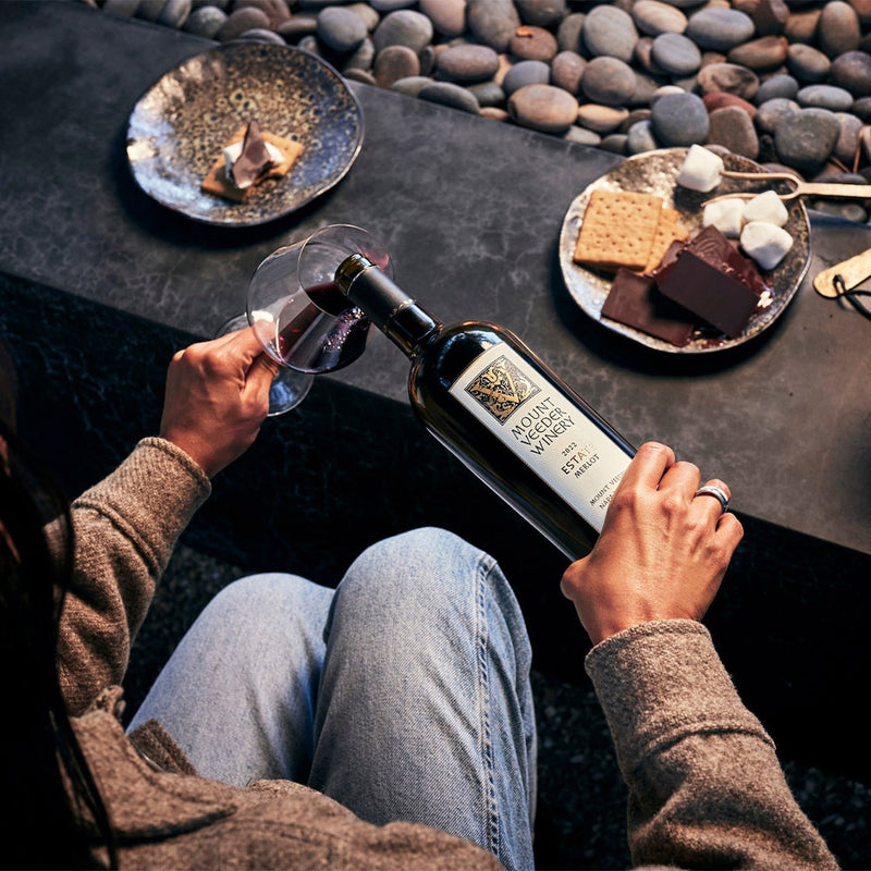 A woman in front of a fire pit pours the Mount Veeder Winery Estate Merlot into a glass with smores on the table 