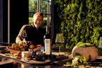 A man at a table with cheese, fruit, a bottle of Mount Veeder Winery Estate Merlot, and a moss wall.