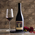 A bottle of The Prisoner Pinot Noir red wine with a cream label and a filled wine glass on a stone surface, styled with red flowers against a textured brown background. 