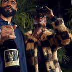 A man in a navy suit holding a bottle of 'The Prisoner' Pinot Noir next to a woman wearing a brown and black checkered faux fur coat, sunglasses, and binoculars against a dark tropical foliage background.