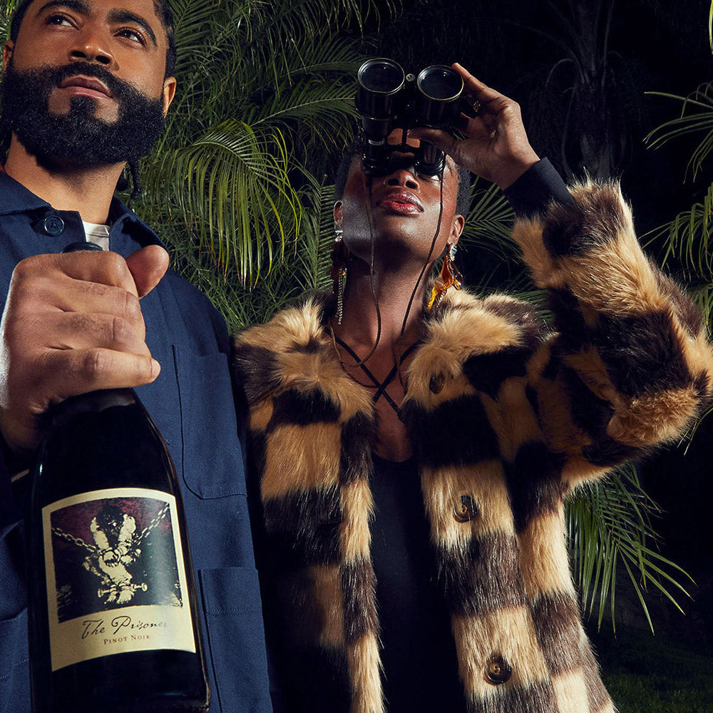 A man in a navy suit holding a bottle of 'The Prisoner' Pinot Noir next to a woman wearing a brown and black checkered faux fur coat, sunglasses, and binoculars against a dark tropical foliage background.